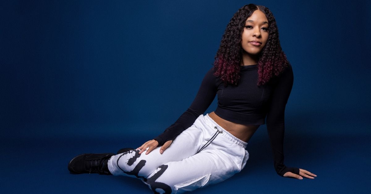 Best Photography Poses For Pictures (From A Pro Photographer) A Woman Poses In A Black Top And White Pants On A Blue Background For A Picture.