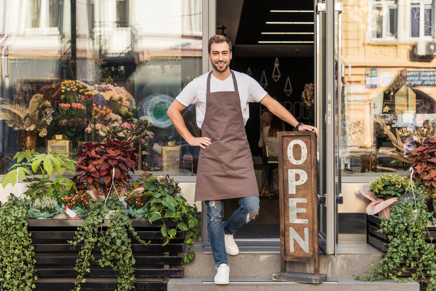 Services A Man Wearing An Apron Stands In The Doorway Of A Flower Shop With Plants And Flowers Around Him And A Sign That Reads &Quot;Open,&Quot; Offering His Services To All Who Enter.