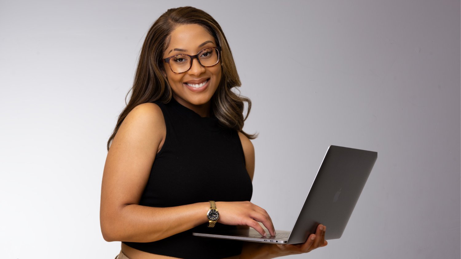 Services A Woman With Glasses, Wearing A Black Sleeveless Top, Smiles At The Camera While Holding A Laptop, Ready To Offer Her Services.
