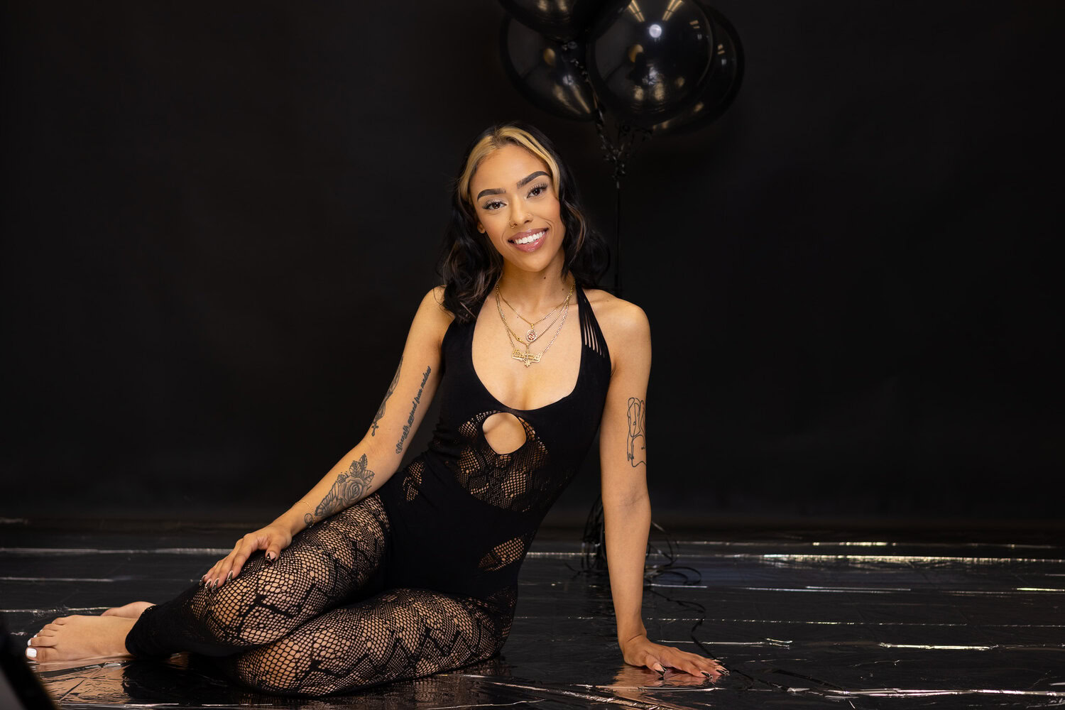Services A Woman With Tattoos, Wearing A Black Patterned Outfit, Sits On The Floor Against A Dark Backdrop With Black Balloons In The Background, Offering Her Unique Services.
