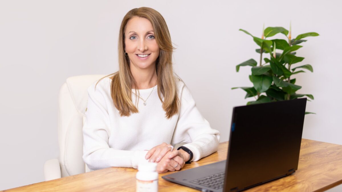 17 Reasons Why Your Business Needs Brand Photography A Woman In A White Sweater Sits At A Desk With A Laptop And A Medication Bottle, Possibly Reviewing Healthcare Services. A Green Plant Is In The Background.
